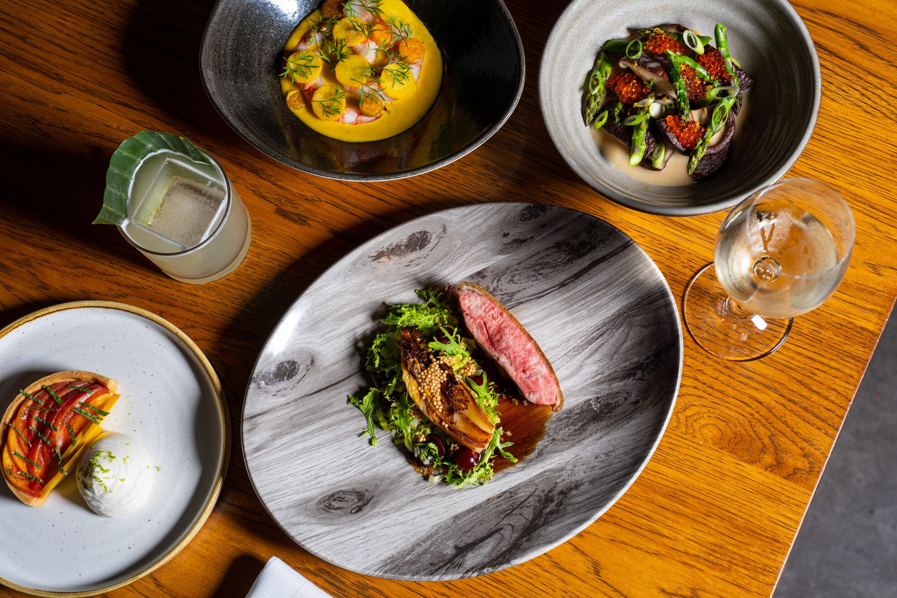 A table is filled with beautifully plated dishes, from seafood and steaks to bright seasonal sides and a craft cocktail. Each plate is styled with fresh herbs, sauces, and vibrant colors. It reflects the creative fine-dining experience Juniper & Ivy is known for.