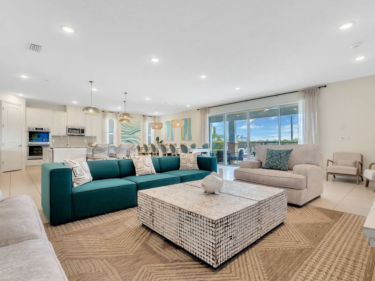 Bright and spacious living area with stylish coastal decor, a deep teal sectional, and sliding glass doors that open to a sunny outdoor space. Perfect for big family groups, the open layout flows easily from the cozy lounge to a full kitchen and long dining table built for memory-making.