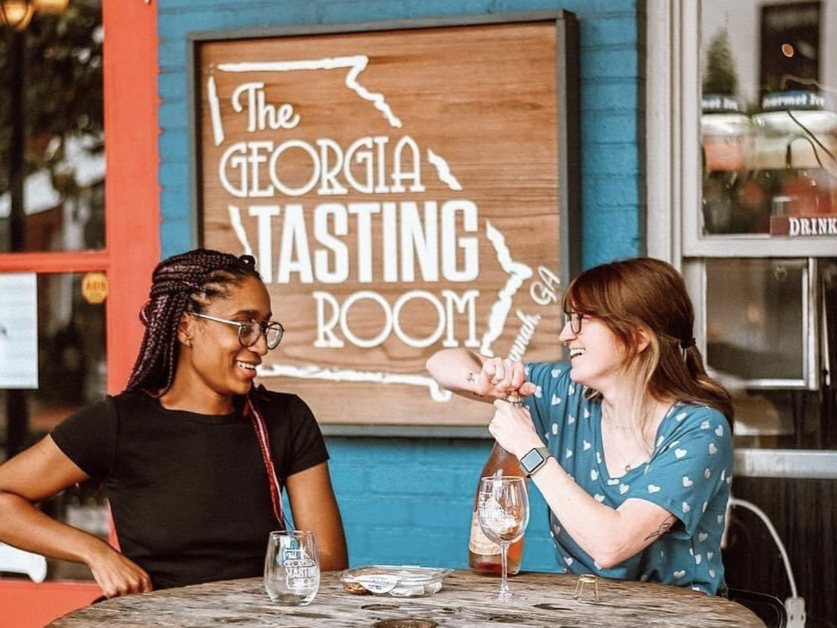 Two friends enjoy a moment at The Georgia Tasting Room, with one opening a bottle of wine as they laugh together at a rustic outdoor table. The setting features a wooden sign on a blue brick wall, adding to the relaxed, local vibe