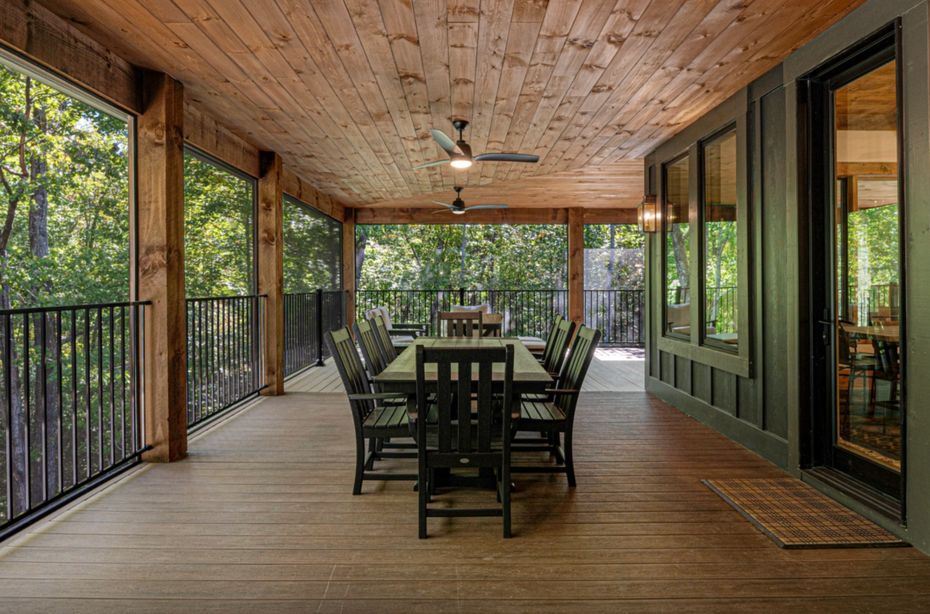 Covered wooden porch with a long dining table and chairs, ceiling fans above, and forest views through screened railings.