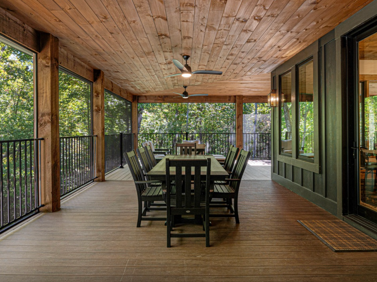 Covered wooden porch with a long dining table and chairs, ceiling fans above, and forest views through screened railings.