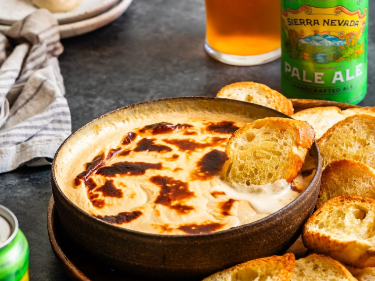 A bowl of baked cheese dip served with toasted bread slices, paired with Sierra Nevada Pale Ale and a glass of beer.