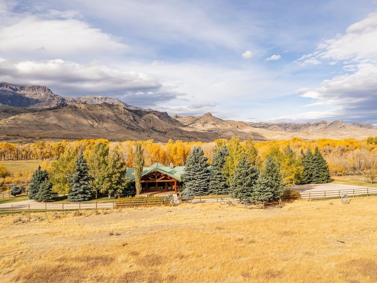 A cozy log cabin with a green roof sits in a valley surrounded by golden autumn trees and distant Wyoming mountains. The wide-open views make this the perfect place to stay for travelers who want nature right outside their door.