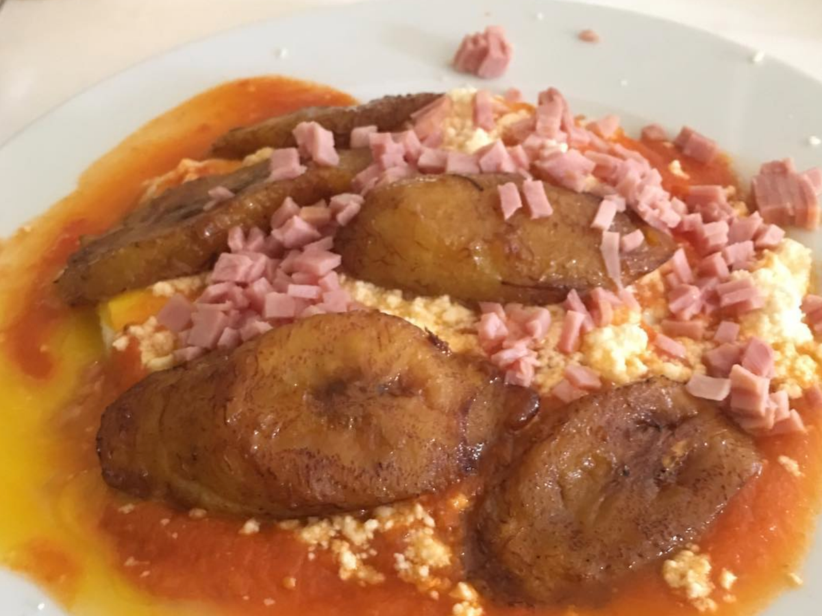 A plate of traditional Mexican enfrijoladas topped with fried plantains, crumbled cheese, diced ham, and a red sauce, creating a rich and savory breakfast dish.
