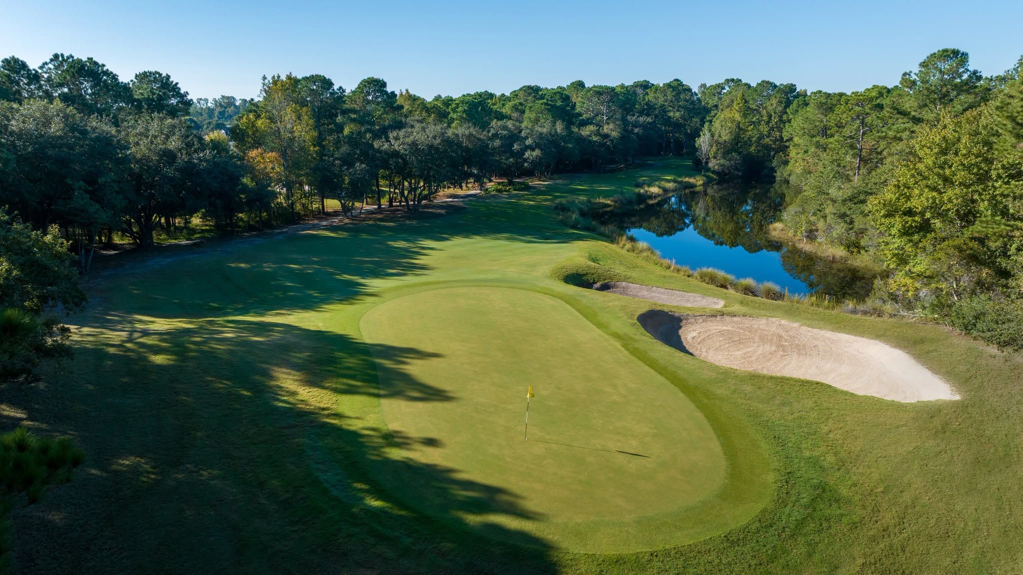A low aerial view of a Barefoot Resort and Golf green in Myrtle Beach features a well-contoured putting surface guarded by two large sand bunkers and a shimmering lake, surrounded by mature oaks and pines in morning light.
