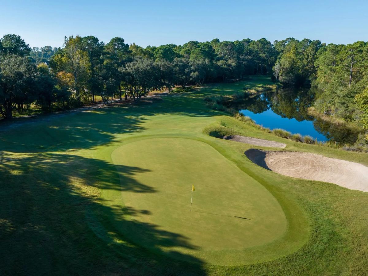 A low aerial view of a Barefoot Resort and Golf green in Myrtle Beach features a well-contoured putting surface guarded by two large sand bunkers and a shimmering lake, surrounded by mature oaks and pines in morning light.