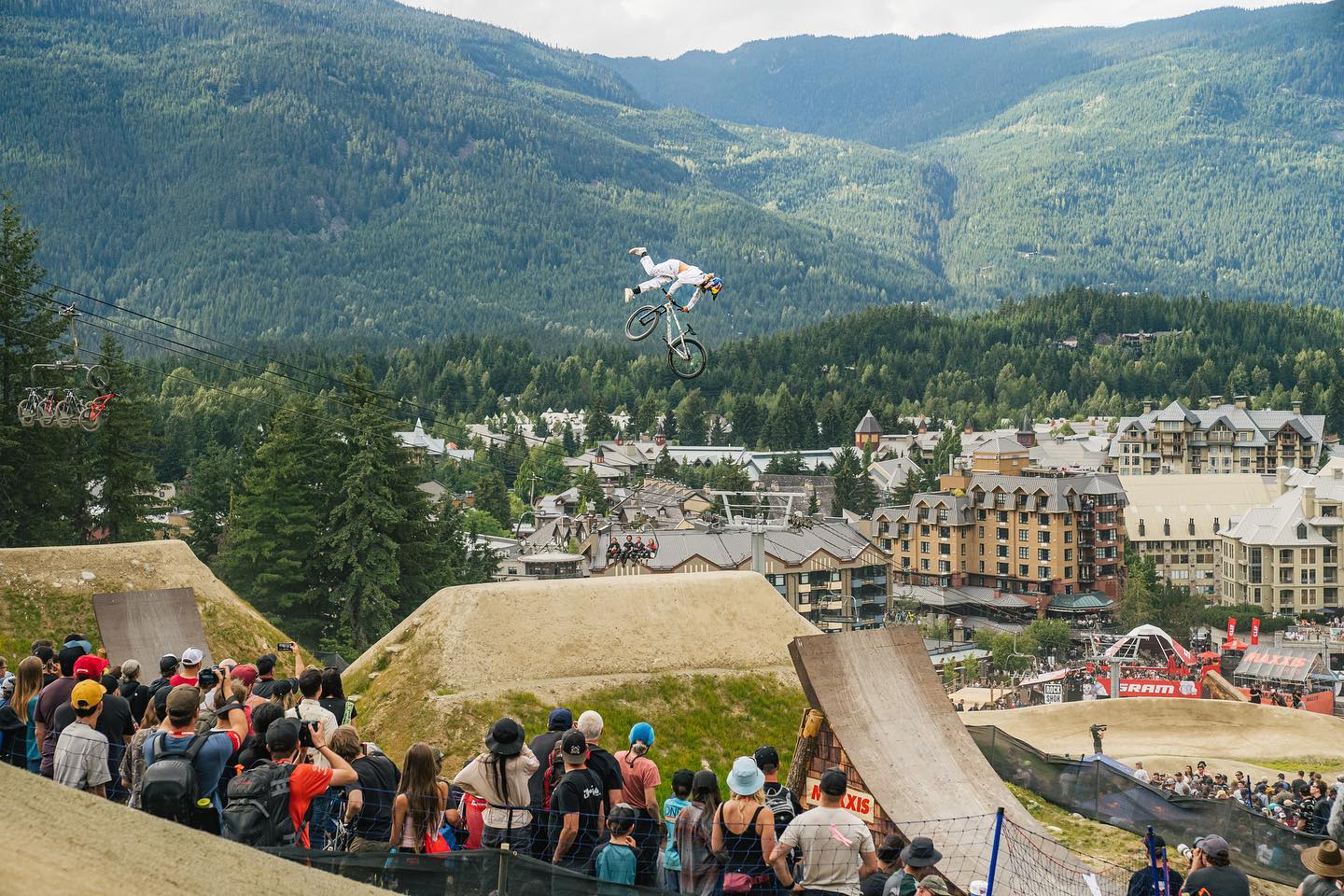 A mountain biker flies high above the dirt jumps at Whistler Mountain Bike Park while a crowd watches below. The village and forested mountains create a dramatic backdrop for summer action. This park is one of the best mountain biking spots in Canada for thrill seekers.