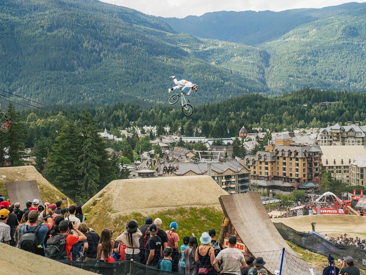 A mountain biker flies high above the dirt jumps at Whistler Mountain Bike Park while a crowd watches below. The village and forested mountains create a dramatic backdrop for summer action. This park is one of the best mountain biking spots in Canada for thrill seekers.