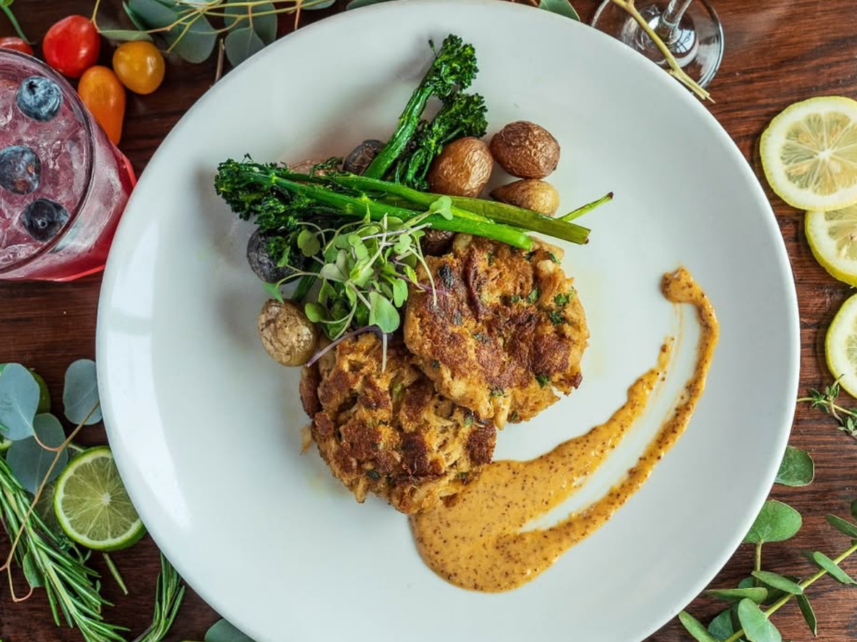 A plated dish of crab cakes served with roasted baby potatoes, broccolini, microgreens, and a smear of mustard aioli, accompanied by a blueberry cocktail and garnished with fresh citrus and herbs.