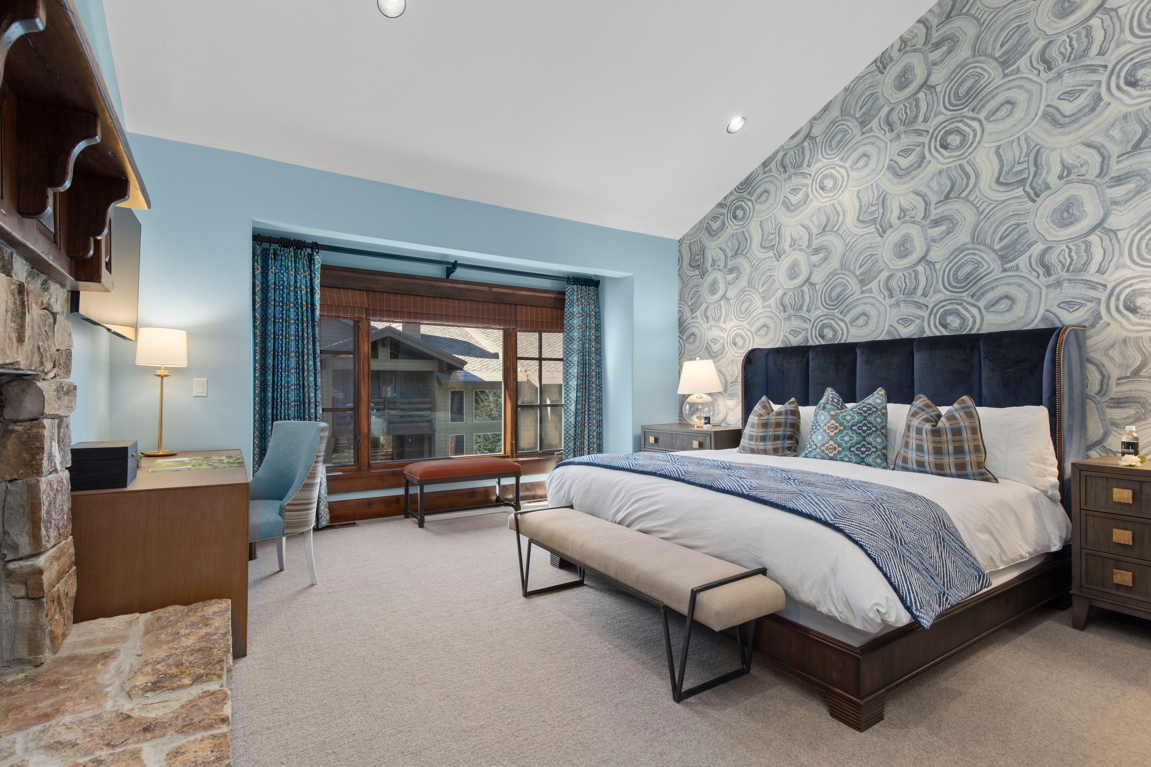 A stylish bedroom with a blue agate-patterned accent wall, a dark upholstered king bed with decorative pillows, a wooden desk near a stone fireplace, and large windows with mountain lodge views.