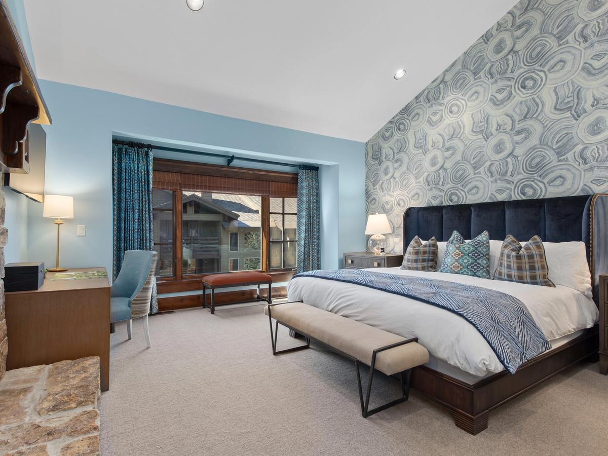 A stylish bedroom with a blue agate-patterned accent wall, a dark upholstered king bed with decorative pillows, a wooden desk near a stone fireplace, and large windows with mountain lodge views.