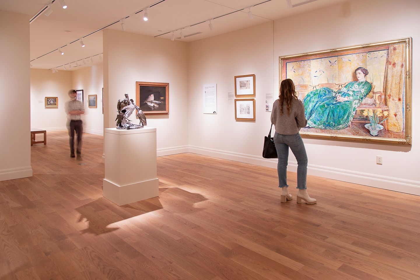 Guests explore a warm, softly lit art gallery with wooden floors and framed paintings lining the walls. A woman pauses in front of a large colorful portrait, taking in the details. The peaceful setting makes it easy to enjoy Charleston’s rich art history.