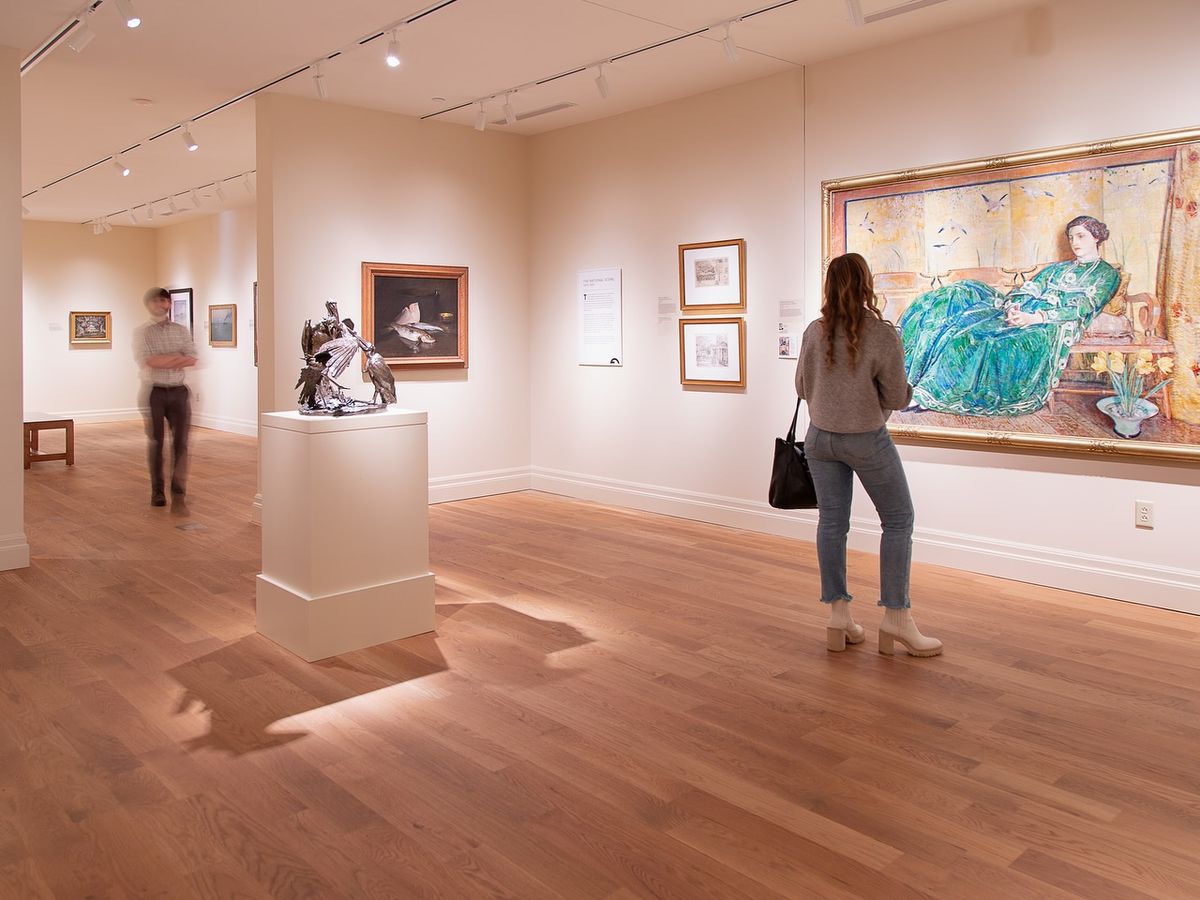 Guests explore a warm, softly lit art gallery with wooden floors and framed paintings lining the walls. A woman pauses in front of a large colorful portrait, taking in the details. The peaceful setting makes it easy to enjoy Charleston’s rich art history.
