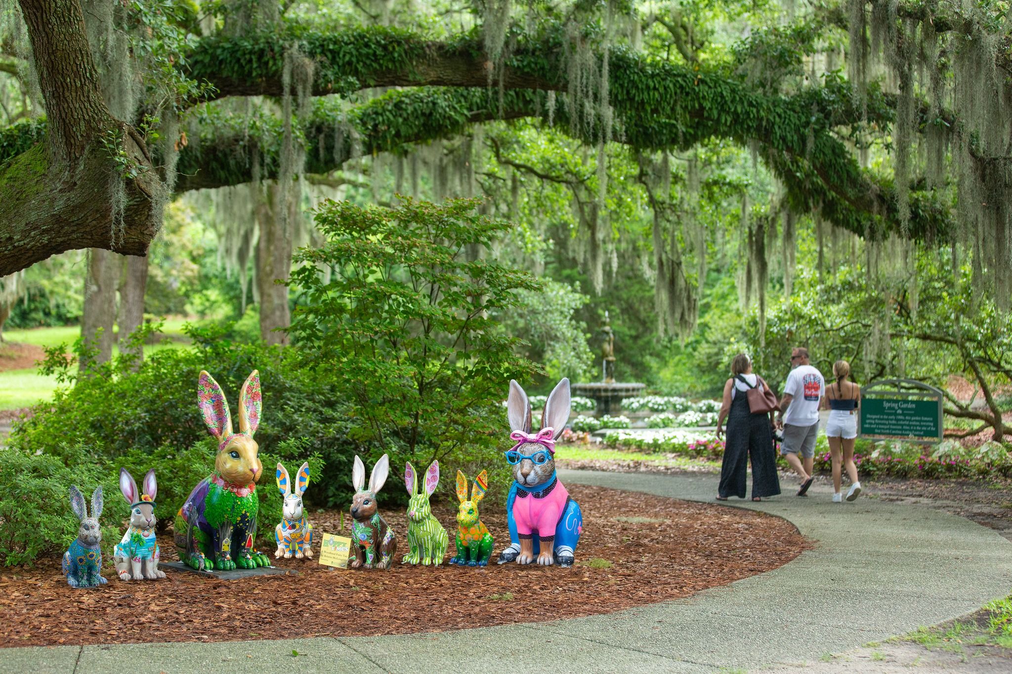 Colorful rabbit statues line a peaceful walking path at Airlie Gardens, surrounded by lush greenery and hanging Spanish moss. Visitors stroll through the garden, enjoying the quiet nature views and unique outdoor art displays.