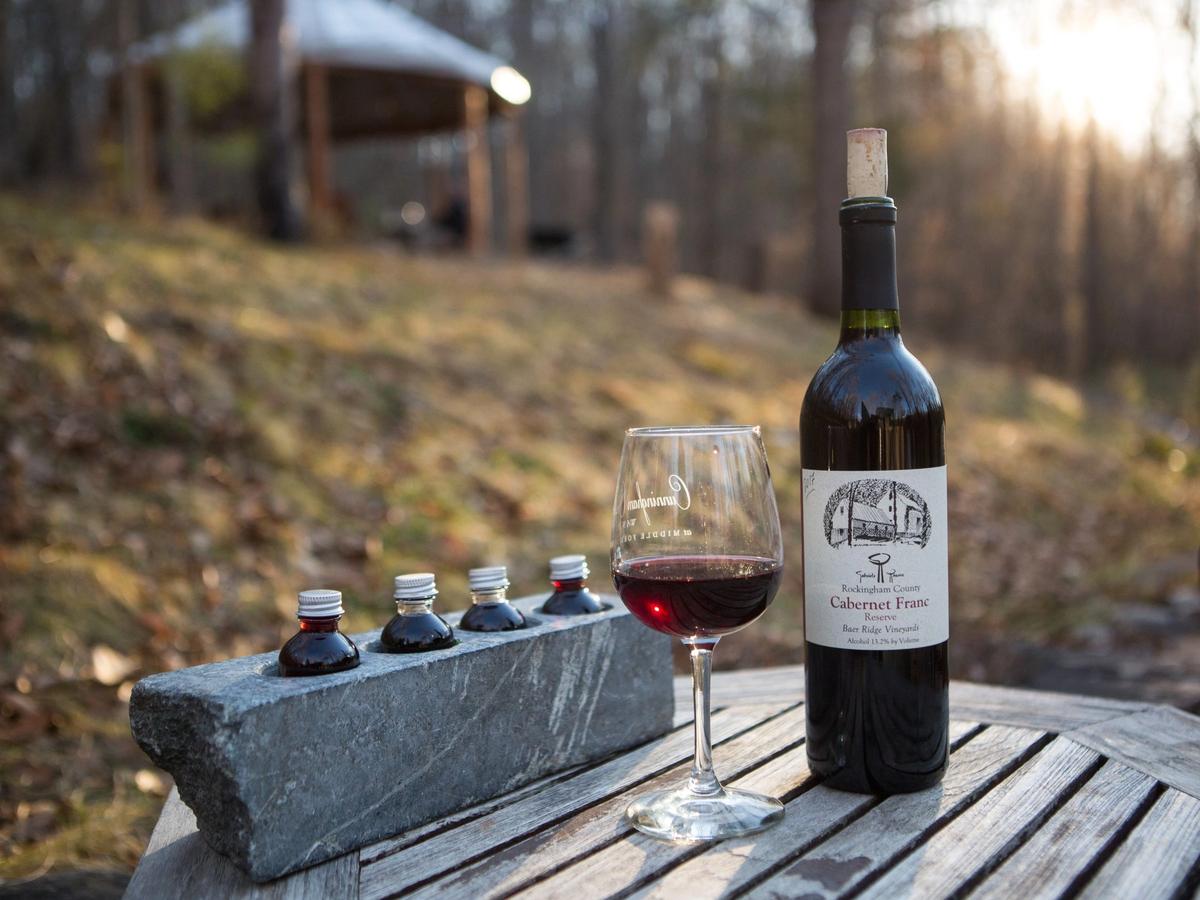 A bottle of Cabernet Franc and a glass of red wine rest on a rustic wooden table outdoors at Gabriele Rausse Winery. The peaceful woodland backdrop and warm sunlight reflect Virginia’s authentic, small-batch wine experience.