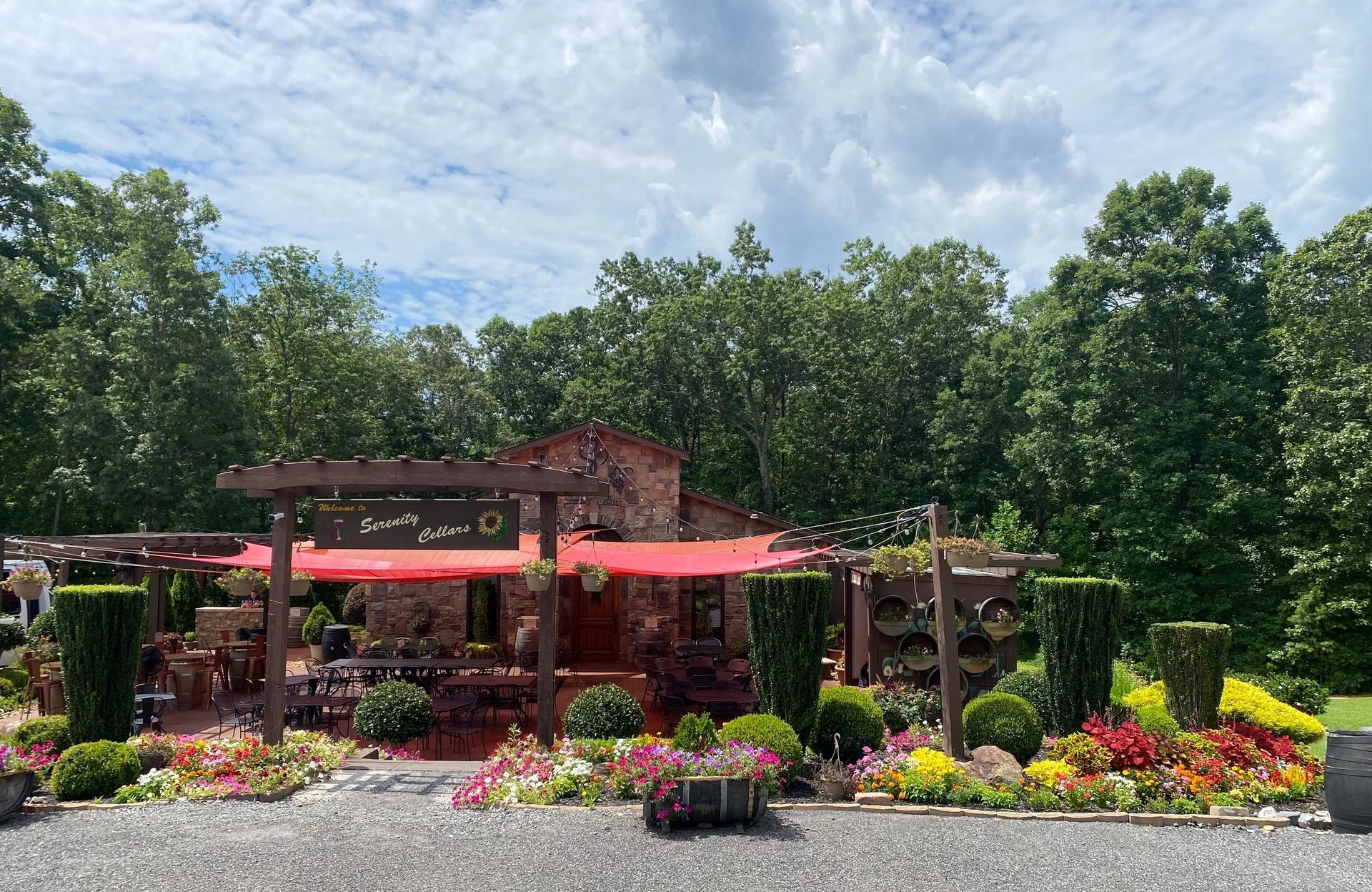 Serenity Cellars features a charming outdoor space filled with colorful flowers, shaded seating, and stone details. The red canopy gives the area a warm, lively feel. It looks like a peaceful place to sit back and enjoy a glass of wine.