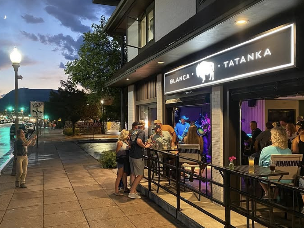 Evening street view of Blanca Tatanka bar with an open-air patio, live music performance inside, and groups of people socializing under a glowing city sky.