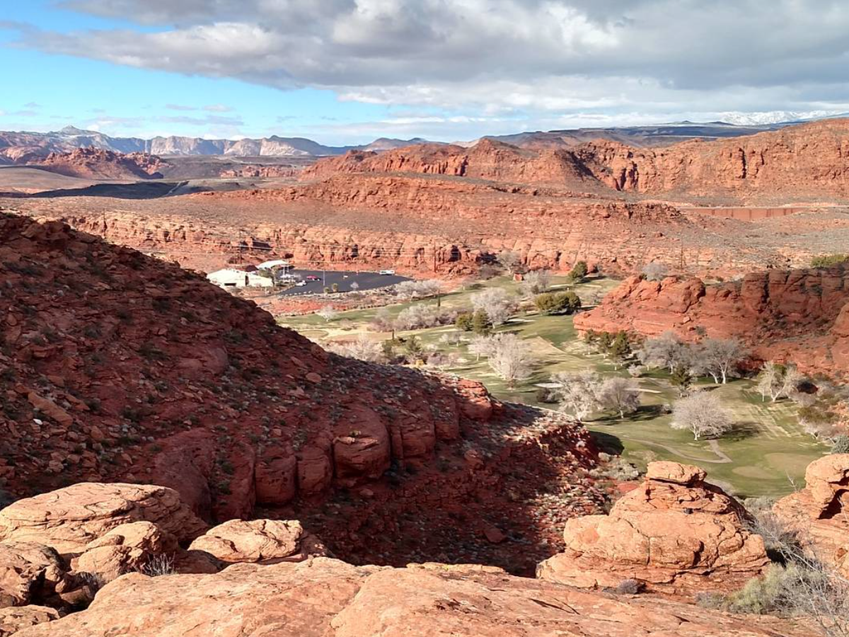 A scenic view of a golf course nestled among red rock canyons, with lush fairways contrasting the rugged desert landscape and distant snow-capped mountains under a partly cloudy sky