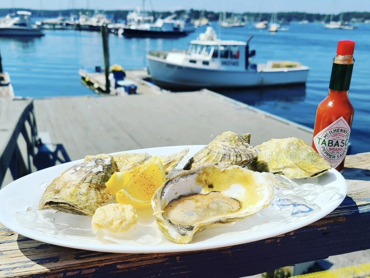 A plate of fresh oysters on the half shell with lemon wedges and horseradish sits on a wooden railing by the harbor, accompanied by a bottle of Tabasco sauce and a scenic view of boats docked in the water.