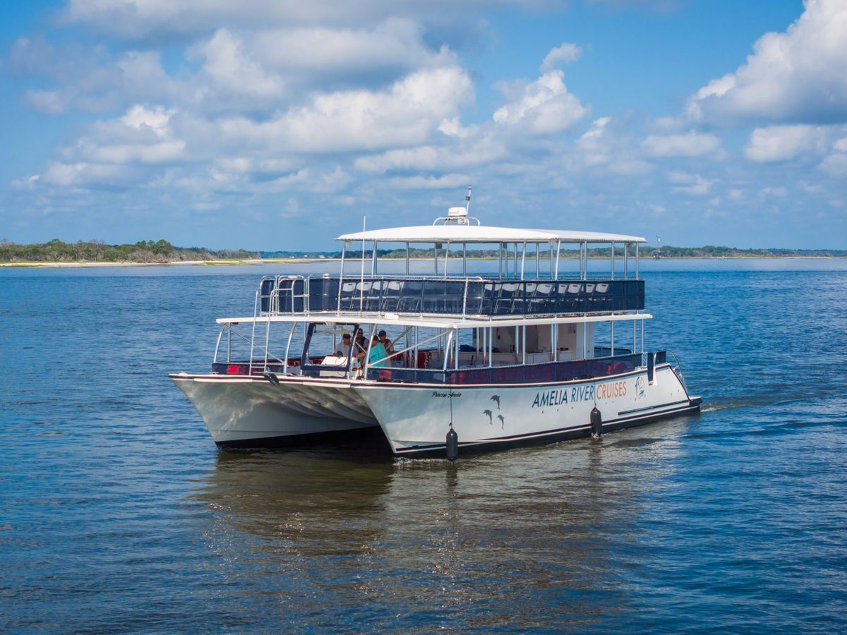 A scenic view of an Amelia River Cruises boat gliding across calm waters under a bright blue sky. The two-level vessel features open-air seating, offering passengers a relaxing and picturesque ride along the coastline
