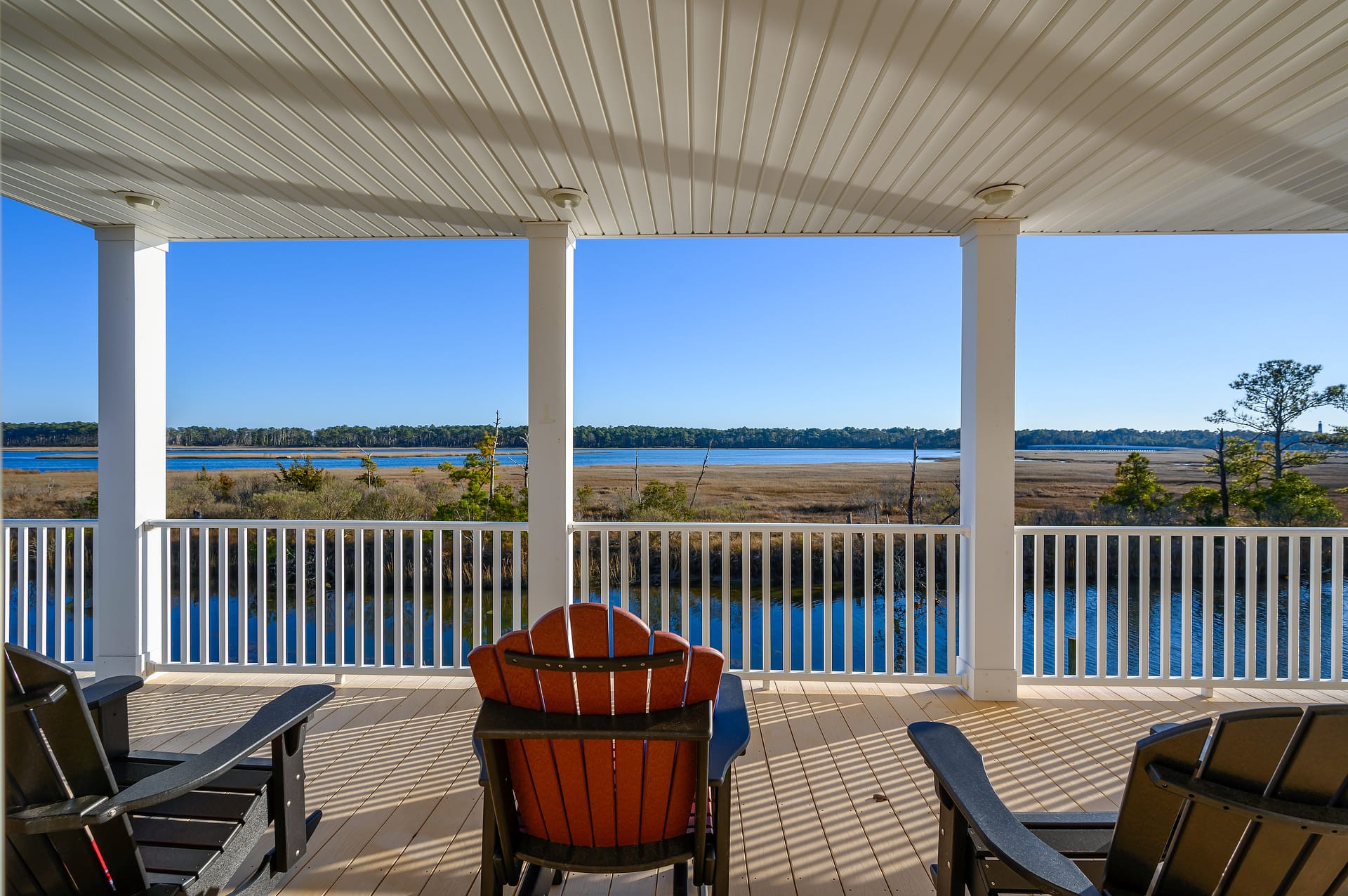 Covered coastal porch with Adirondack chairs overlooking a marsh and calm blue water framed by white columns under a beadboard ceiling.