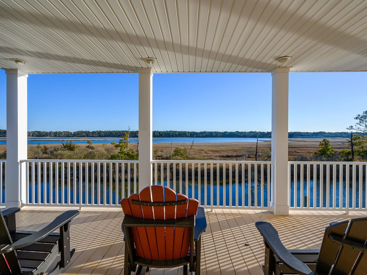 Covered coastal porch with Adirondack chairs overlooking a marsh and calm blue water framed by white columns under a beadboard ceiling.