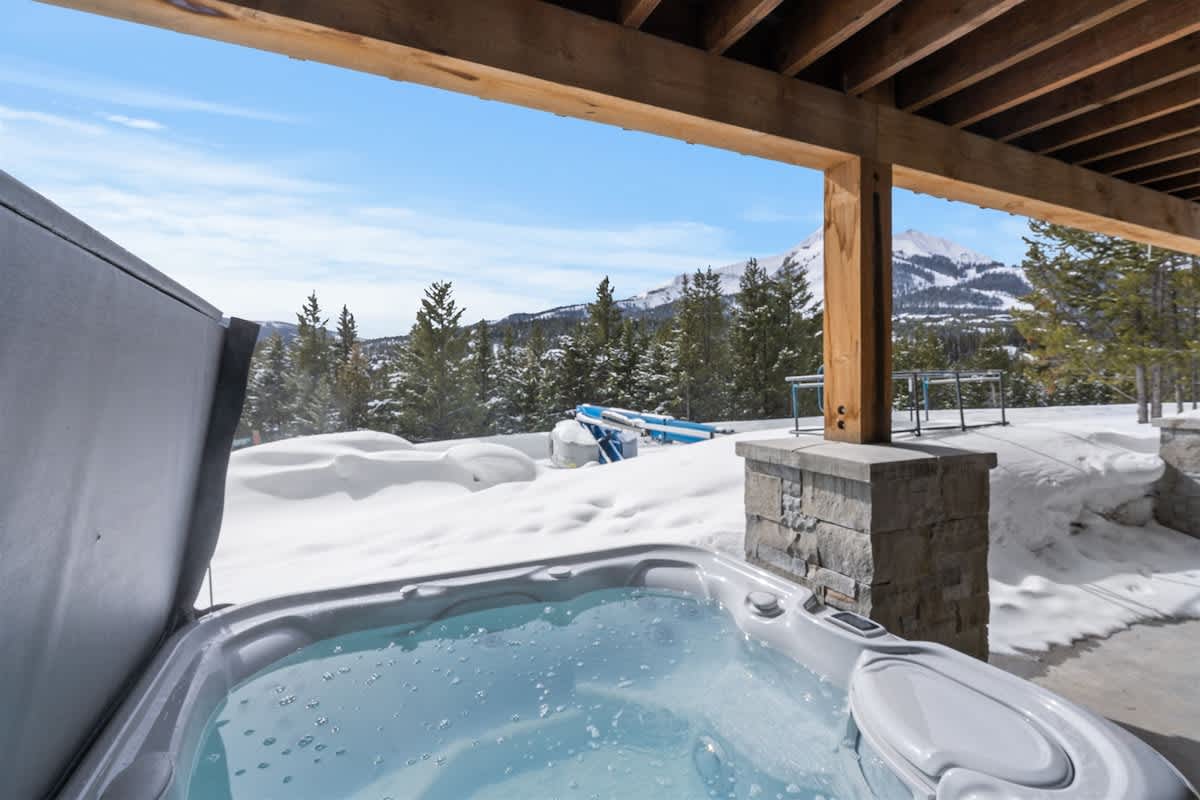 Covered outdoor hot tub with clear, steaming water on a mountain cabin patio, surrounded by snow-covered pine trees and distant alpine peaks, offering a peaceful winter soak in a scenic high-elevation setting.