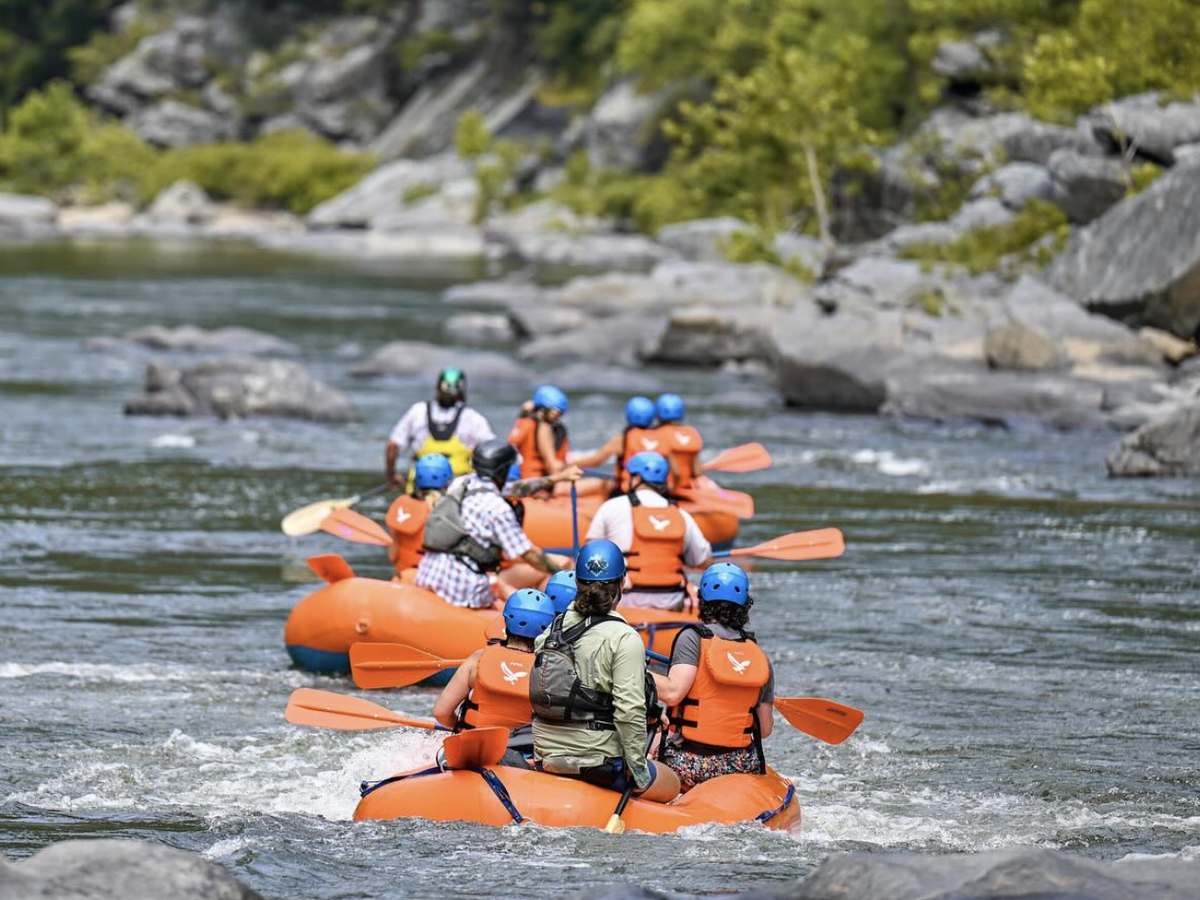 Groups of people wearing life jackets and helmets paddle orange rafts down a scenic, rocky river surrounded by greenery.