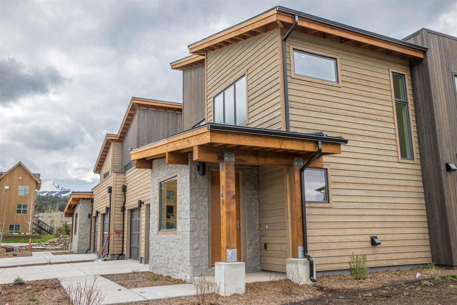 A modern mountain townhouse with wood siding, stone accents, and a covered entryway sits under a cloudy sky, with snow-capped peaks visible in the background.