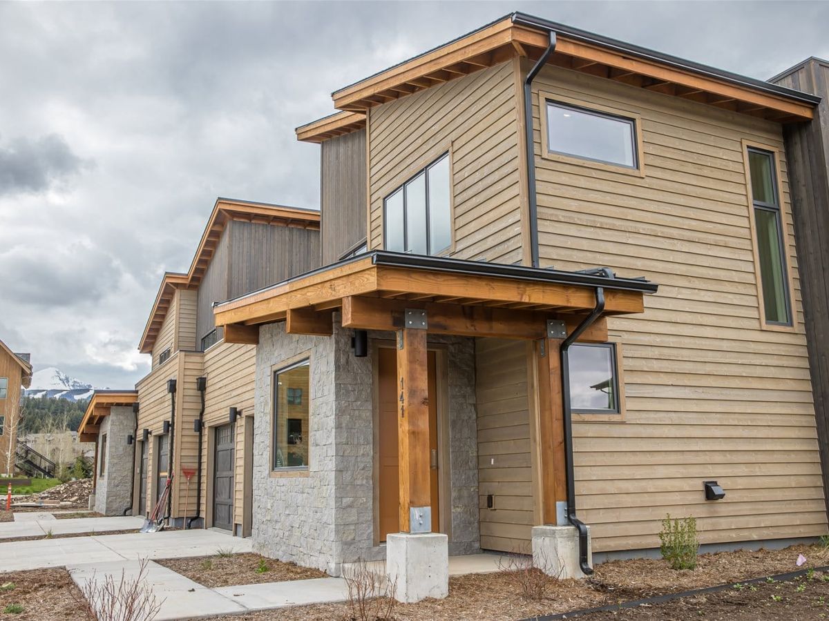 A modern mountain townhouse with wood siding, stone accents, and a covered entryway sits under a cloudy sky, with snow-capped peaks visible in the background.