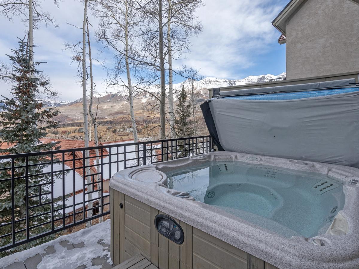 A private hot tub on a snowy balcony overlooks a scenic mountain landscape, framed by aspen trees and rooftops with snow-dusted red tiles.