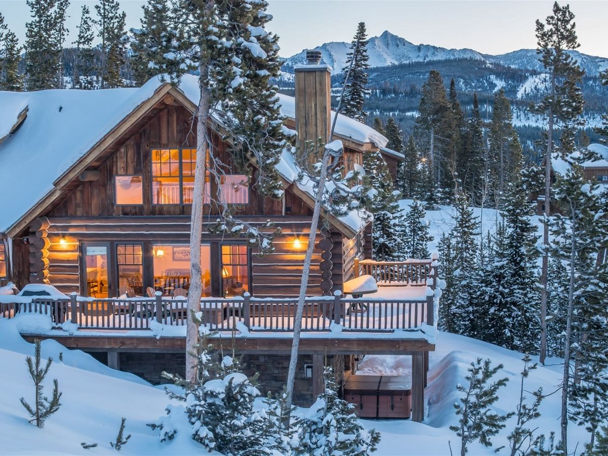 A cozy log cabin glows warmly at sunset, surrounded by fresh snow and tall pine trees in Big Sky. With mountain views in the distance, it’s the perfect winter getaway for a peaceful and private escape.