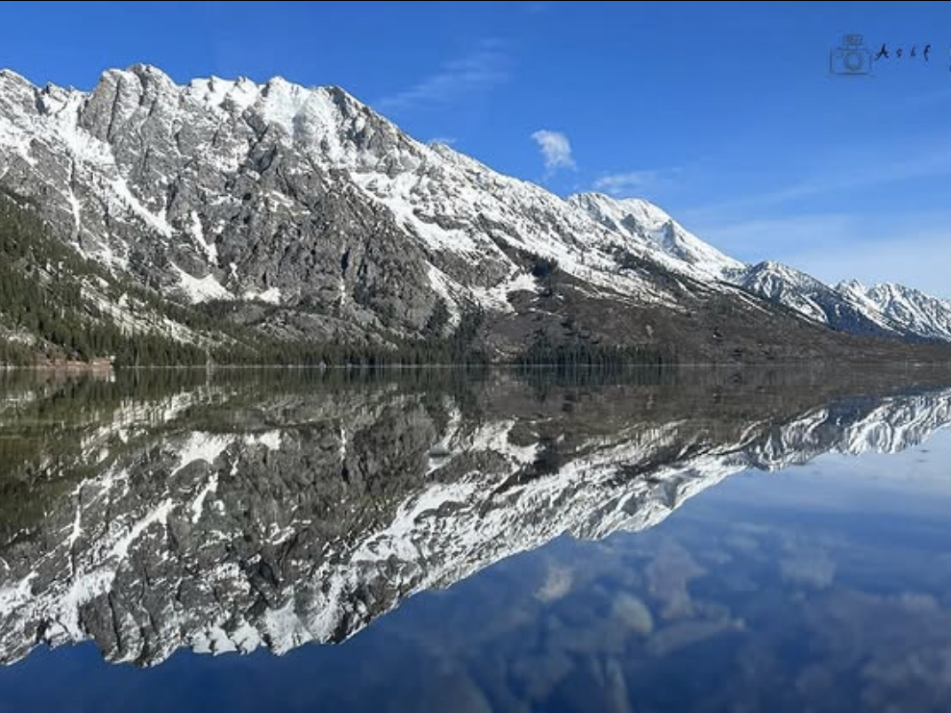 Snow-covered mountain range reflected perfectly in a still, clear lake under a bright blue sky.