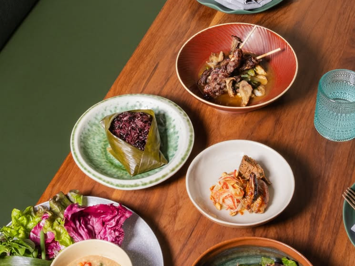A vibrant spread of Southeast Asian-inspired dishes including grilled meat skewers, purple sticky rice wrapped in banana leaf, fresh greens with dipping sauce, papaya salad with chicken, and a colorful vegetable stir-fry, arranged on a wooden table with modern tableware.