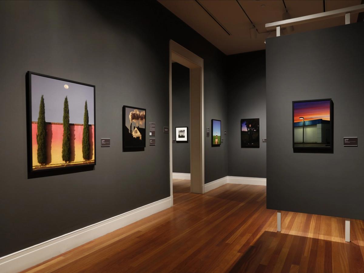 This gallery room features bold, colorful photographs displayed on deep gray walls, creating a striking contrast with the warm wooden floors. Each piece shows a different scene, from tall trees against a sunset wall to glowing nighttime landscapes. The quiet space feels perfect for exploring modern Southern art at your own pace.