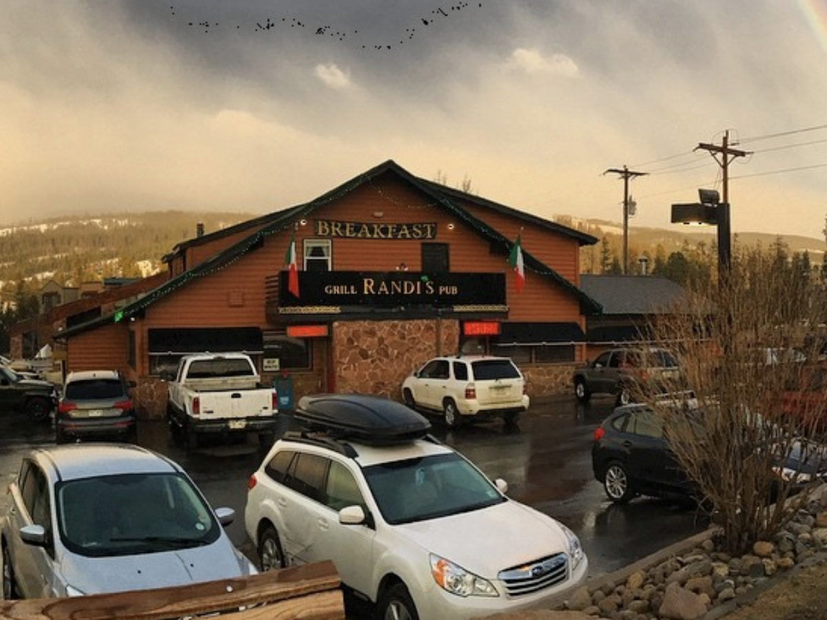Rustic mountain pub called Randi’s Grill & Pub with a stone and wood exterior, parked cars in front, and a rainbow arcing across the cloudy sky above snowy hills.