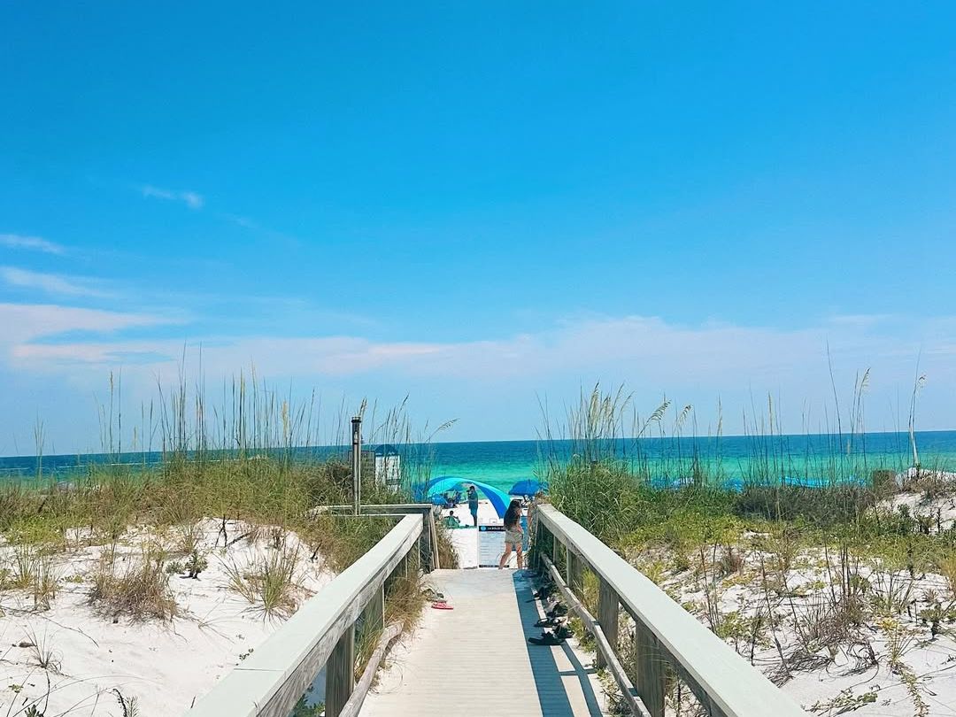 A bright summer day at Okaloosa Island with a wooden boardwalk leading to soft white sand and emerald-green Gulf waters, perfect for a beach getaway.