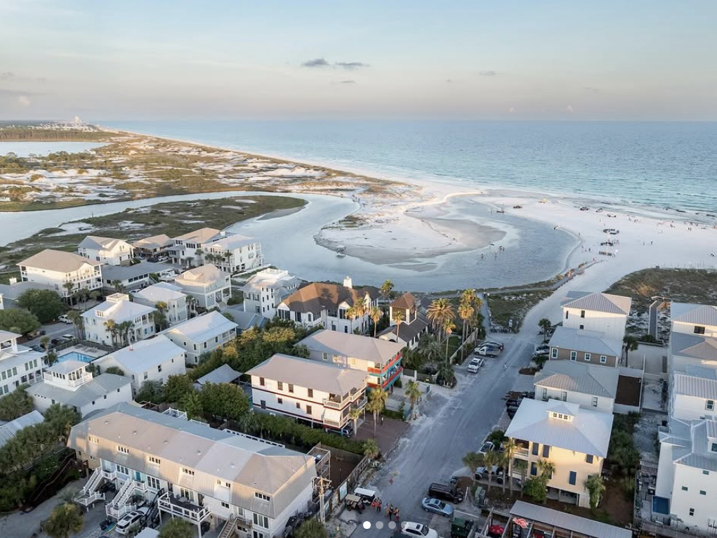 Aerial view of a coastal neighborhood with white-roofed houses, palm trees, and cars parked along the streets, leading out to a wide sandy beach and calm turquoise ocean waters at sunset.