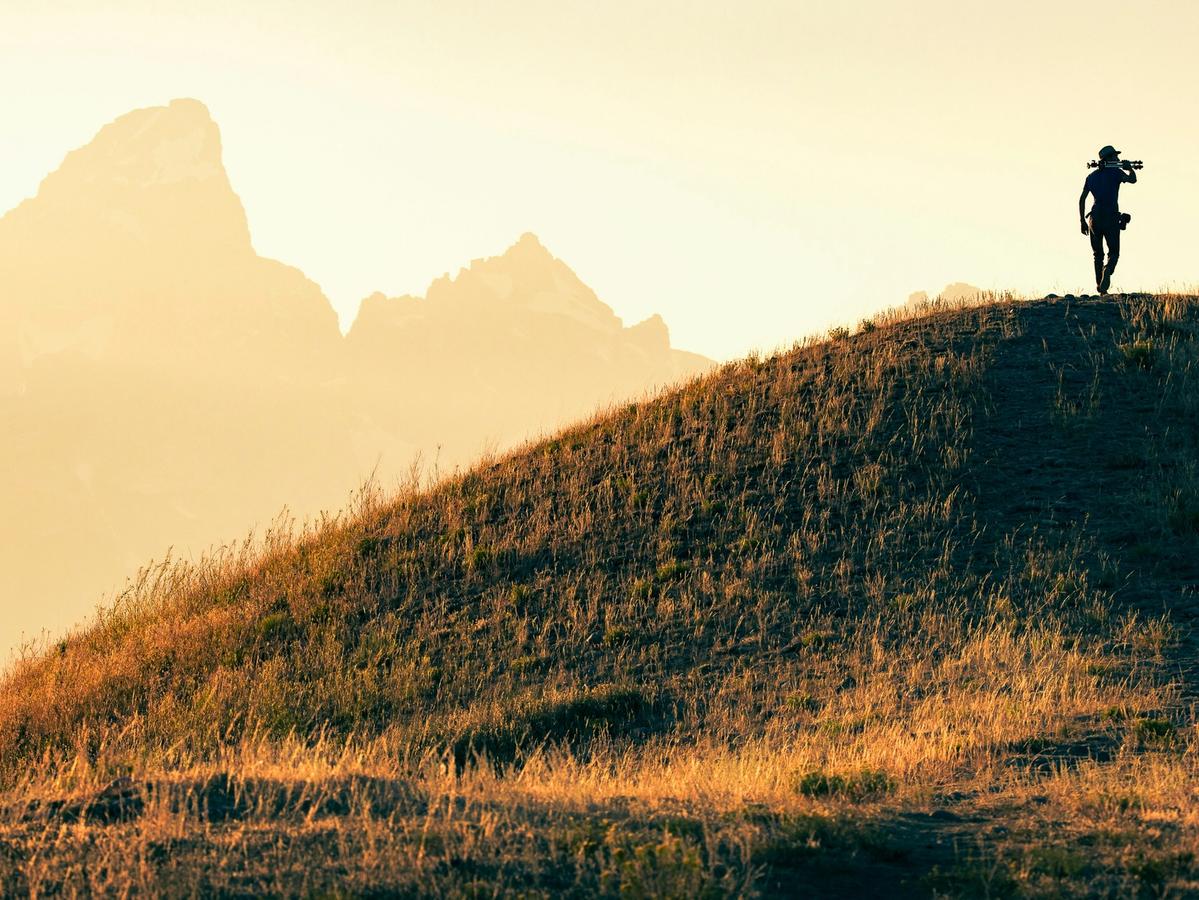 A lone hiker silhouetted against the golden light of dawn walks along a grassy ridge, with the majestic peaks of the Tetons rising softly in the misty background.