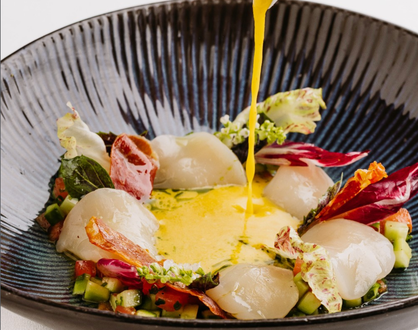 Elegant seafood dish featuring tender scallops arranged over finely diced vegetables and crisp garnishes, finished with a vibrant golden sauce being poured tableside.