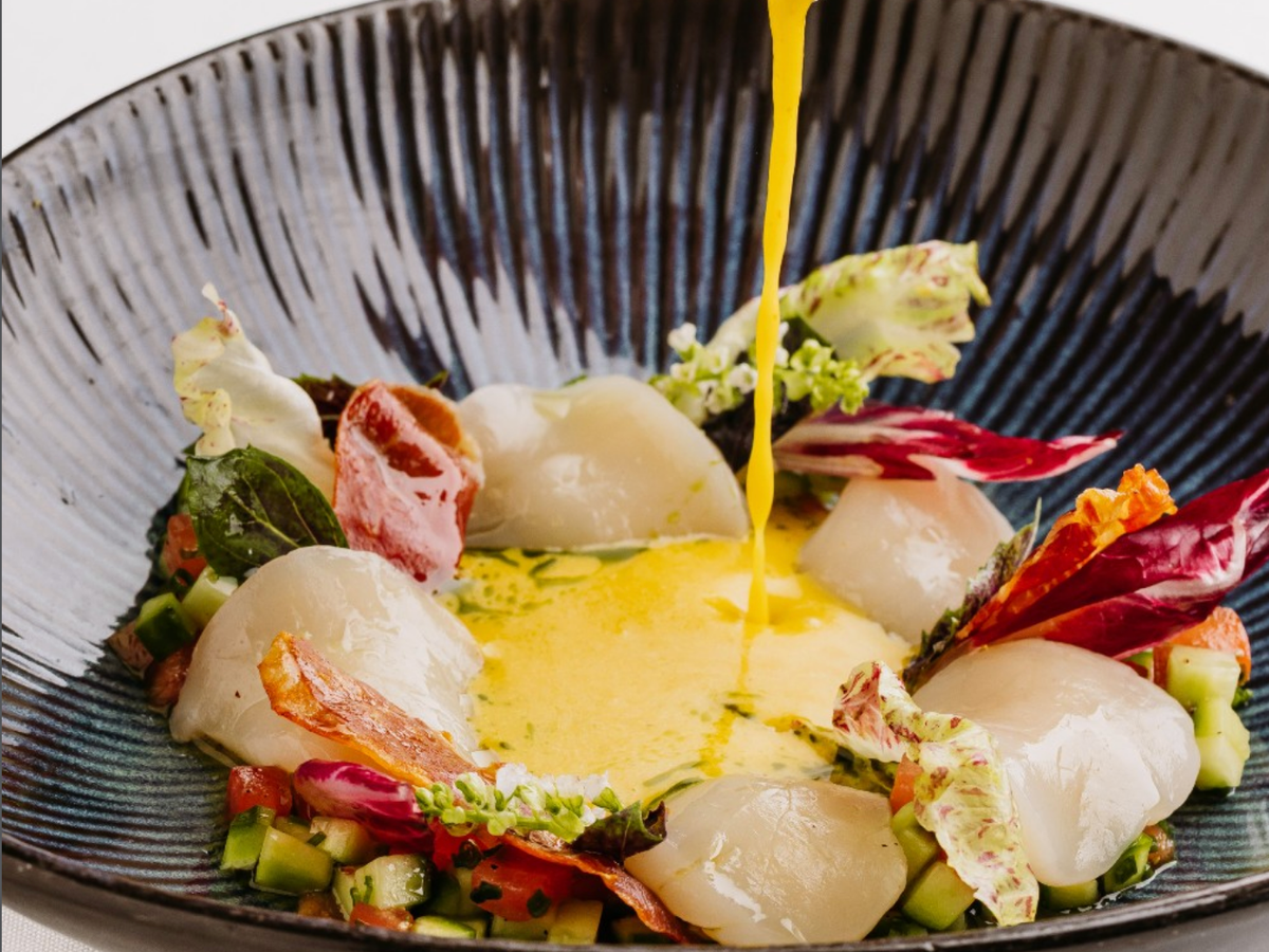 Elegant seafood dish featuring tender scallops arranged over finely diced vegetables and crisp garnishes, finished with a vibrant golden sauce being poured tableside.