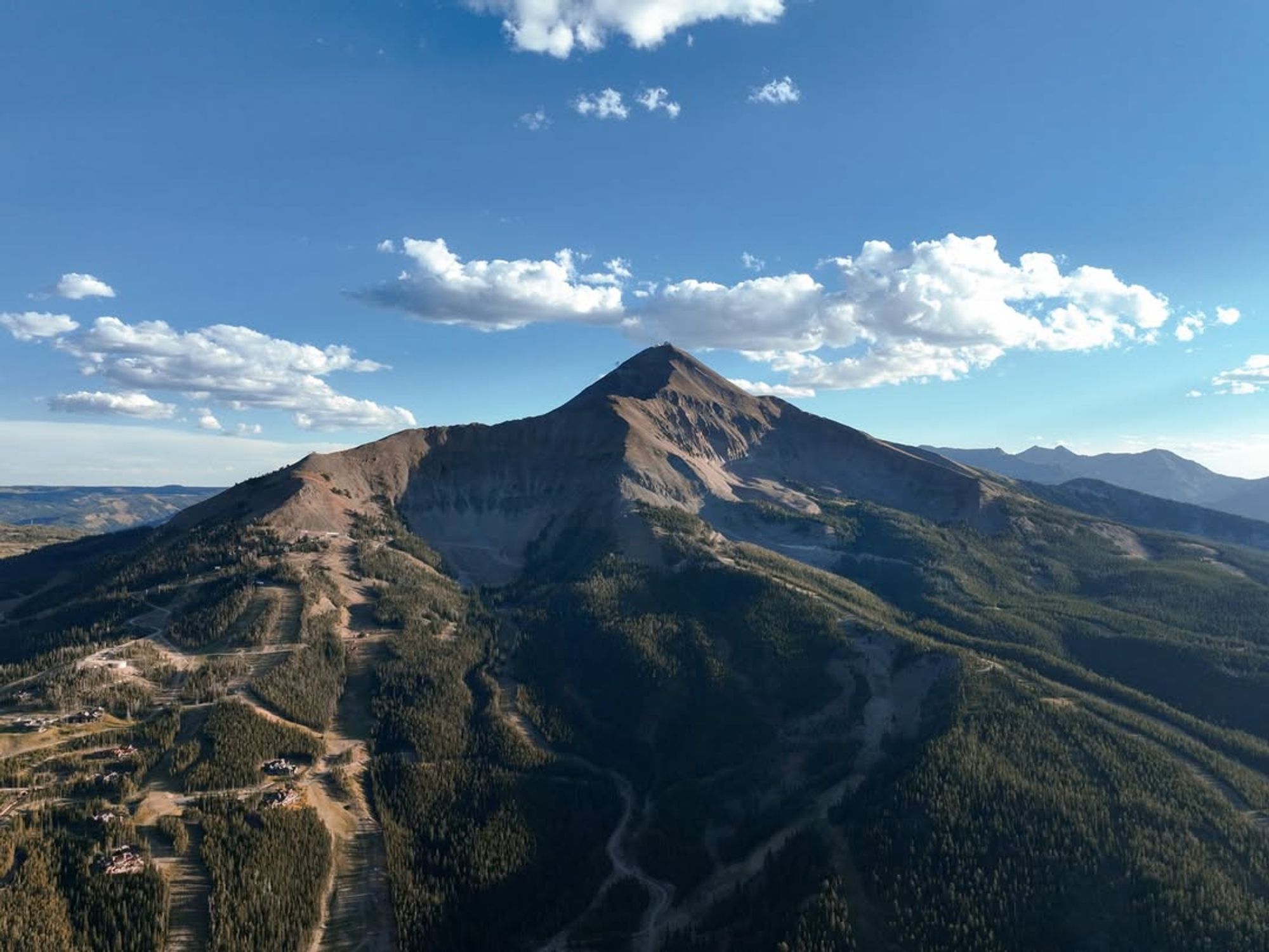  A stunning aerial view of Lone Peak towers over pine-covered slopes and winding trails. Big Sky’s iconic mountain stands tall against a backdrop of blue skies and scattered clouds.