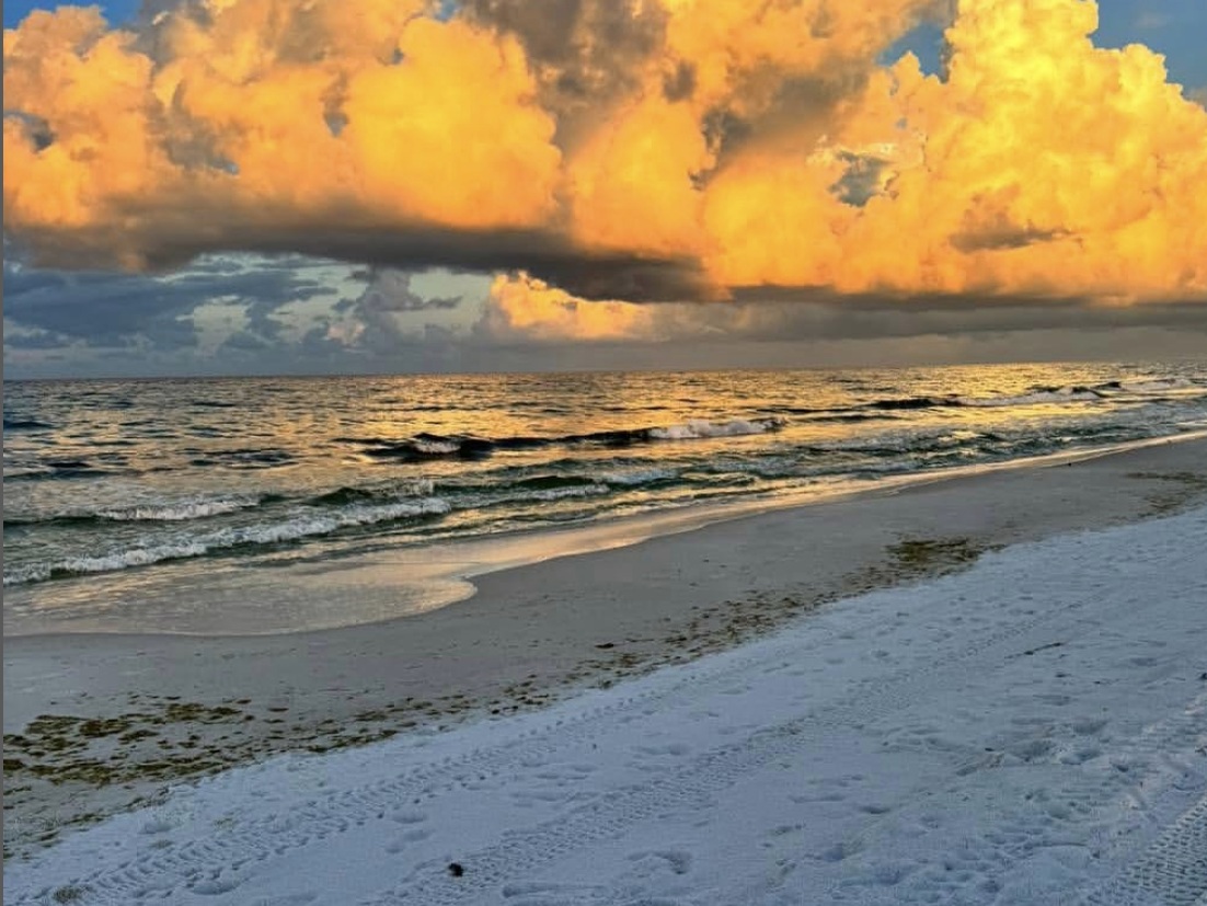 Golden sunset clouds glow dramatically over the ocean, reflecting on the rippling waves, while the white sandy beach stretches forward with footprints and tire tracks.