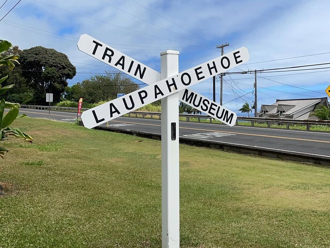 A white signpost with arrows pointing to "Train," "Laupahoehoe," and "Museum" stands on green grass near a quiet roadside. This small-town museum offers a peek into Hawaii’s railroad history.