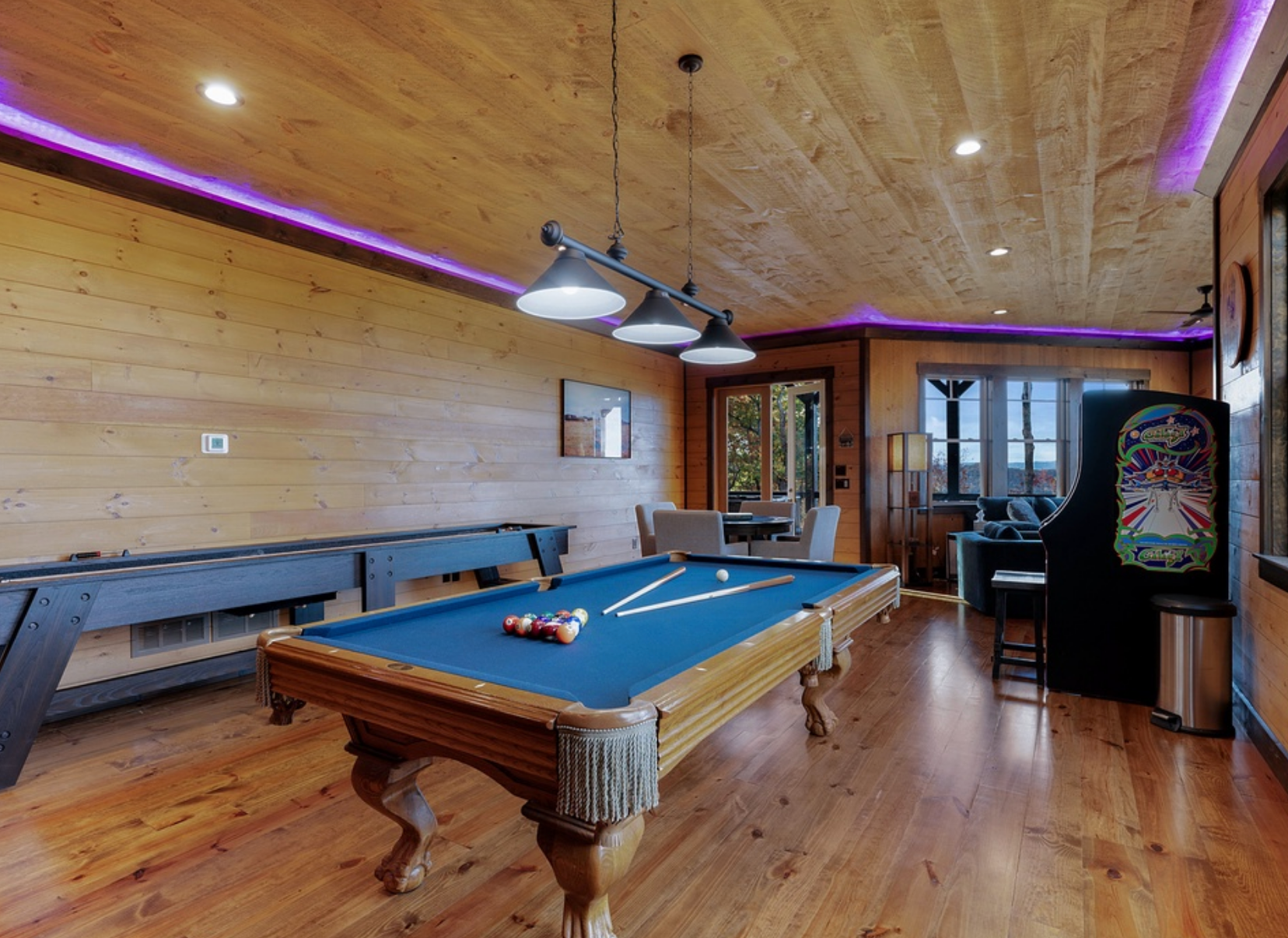 A warm wood-paneled game room features a classic pool table under industrial pendant lights, with cues and balls set for play. Purple LED accent lighting lines the ceiling, while an arcade machine and cozy seating area sit near large windows overlooking scenic views.