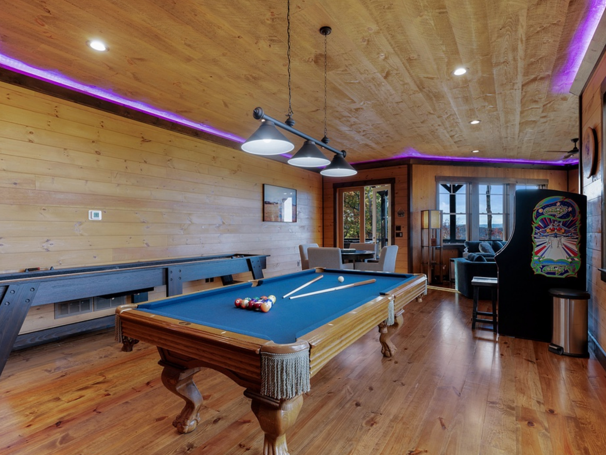A warm wood-paneled game room features a classic pool table under industrial pendant lights, with cues and balls set for play. Purple LED accent lighting lines the ceiling, while an arcade machine and cozy seating area sit near large windows overlooking scenic views.