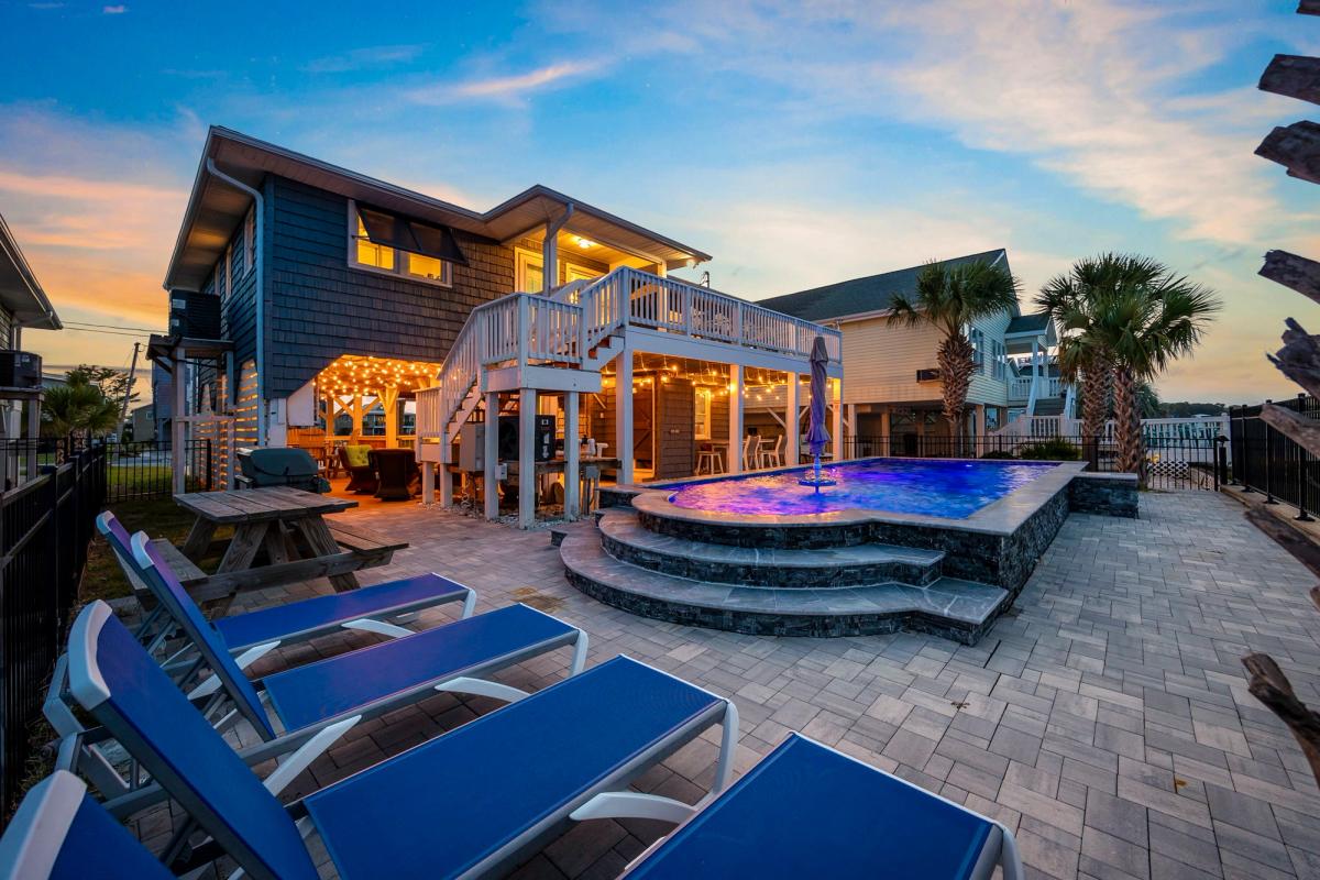This cozy waterfront home shines at sunset, with string lights twinkling around the pool deck and lounge chairs ready for evening relaxation. The elevated deck and outdoor bar area make it perfect for family BBQs and gatherings after a beach day.