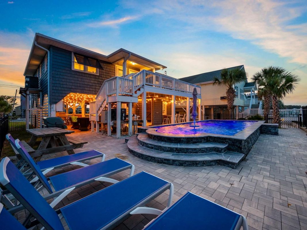 This cozy waterfront home shines at sunset, with string lights twinkling around the pool deck and lounge chairs ready for evening relaxation. The elevated deck and outdoor bar area make it perfect for family BBQs and gatherings after a beach day.