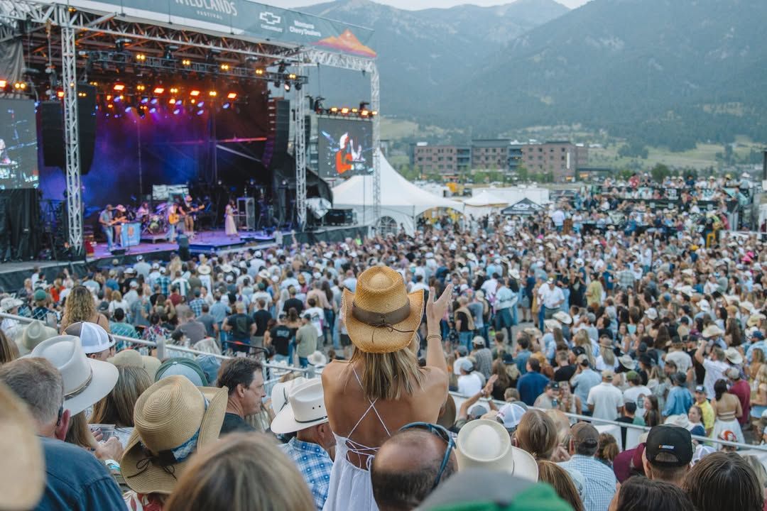 A packed outdoor concert lights up the evening as fans in cowboy hats dance and cheer to live country music. The mountain scenery and bright stage lights add to the energy of the summer festival.