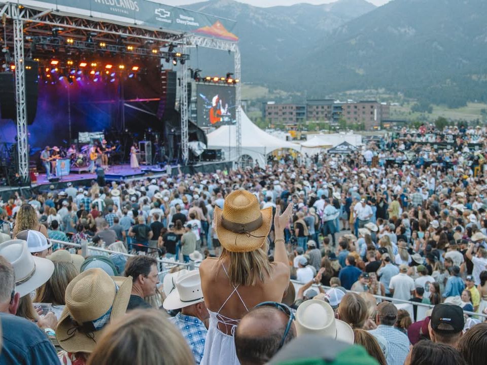 A packed outdoor concert lights up the evening as fans in cowboy hats dance and cheer to live country music. The mountain scenery and bright stage lights add to the energy of the summer festival.